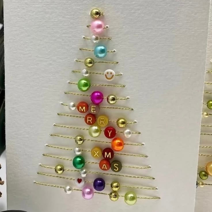 A handmade Christmas greeting card featuring a gold wire tree outline decorated with colorful beads and letter tiles spelling 'MERRY CHRISTMAS' on a textured white cardstock.