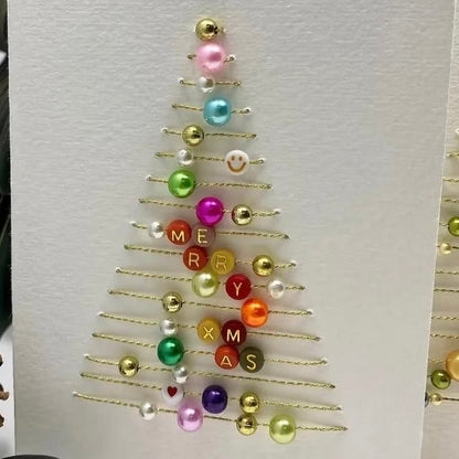 A handmade Christmas greeting card featuring a gold wire tree outline decorated with colorful beads and letter tiles spelling 'MERRY CHRISTMAS' on a textured white cardstock.