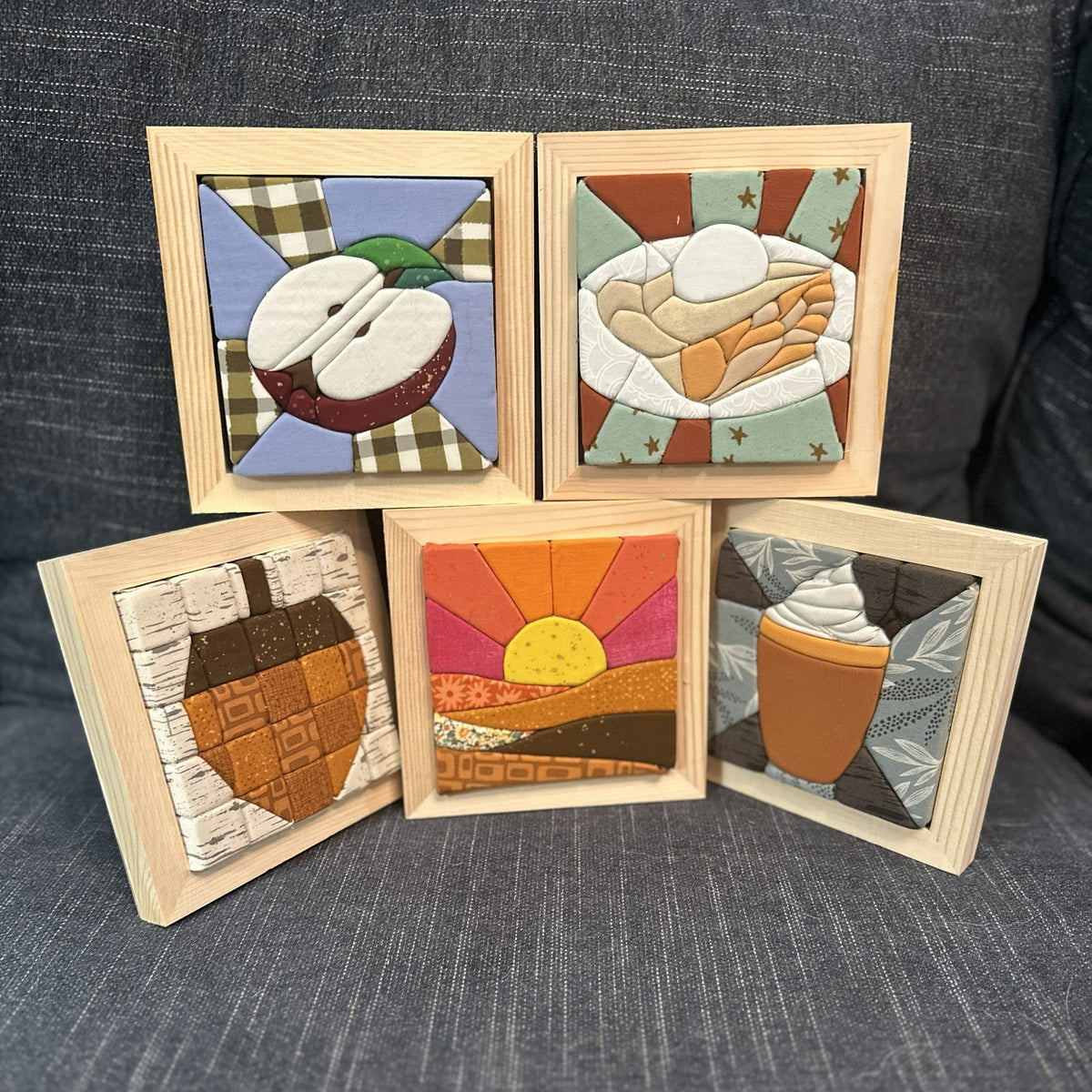 Five small square framed art pieces with light wood frames are arranged on a dark gray textured fabric surface. Each piece features a different illustrated scene: a sliced apple, pastries, an acorn, a sunset, and a hot drink with whipped topping.