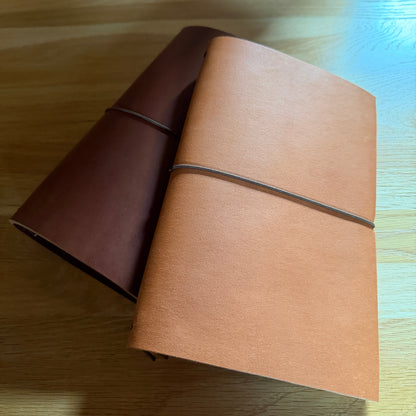 Two leather journal covers or notebooks with elastic closures, one in dark brown and one in light tan, resting on a light wood tabletop.