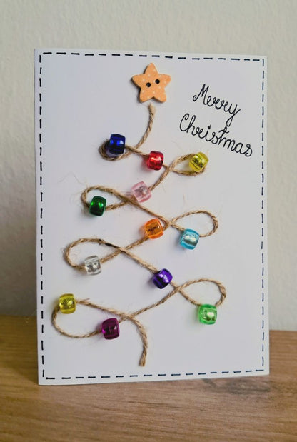 A handmade Christmas greeting card featuring a white background with a dashed black border, decorated with twisted jute rope forming a tree shape and small colorful glass beads. A wooden star ornament hangs from the top, and the words 'Merry Christmas' are written in black script on the right.