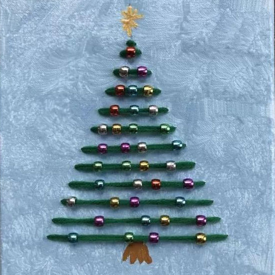 A handmade Christmas tree decoration made from green yarn stripes and shiny multicolored beads, topped with a gold star and finished with a gold bow at the bottom, set against a light blue textured fabric background.